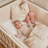 Baby in a cream FLEXA baby bed, 140x70 cm, with beige blanket and plush octopus, shown in a nursery setting.