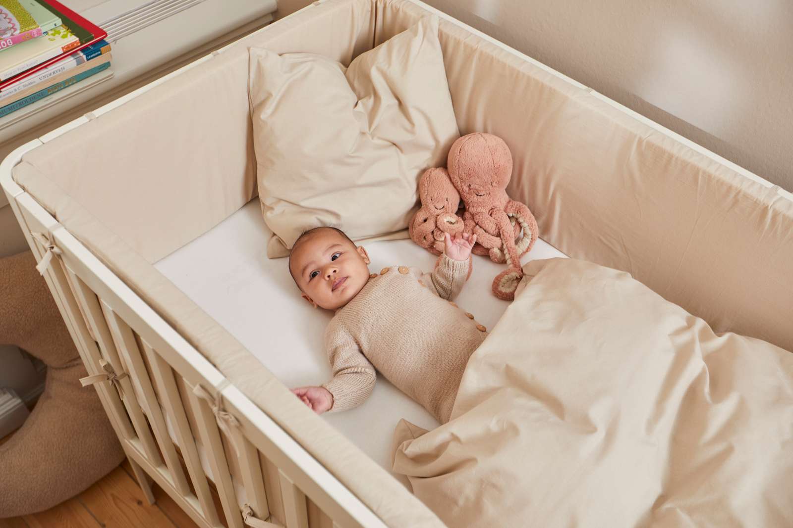 Baby in a cream FLEXA baby bed, 140x70 cm, with beige blanket and plush octopus, shown in a nursery setting.