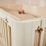 FLEXA baby bed, 140x70 cm, cream finish, visible soft padding, child resting inside; nursery context.