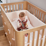 Baby with pacifier in FLEXA baby bed, OAK, soft bedding and bumpers, nursery; child-safe rails visible.