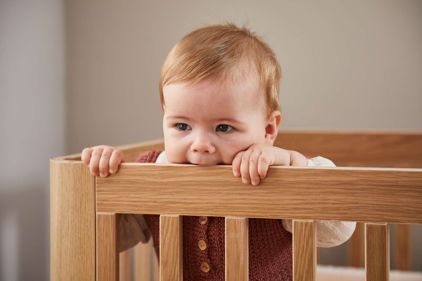 Baby in FLEXA OAK cot bed, safely biting top rail; child-safe design, oak finish, nursery setting visible.
