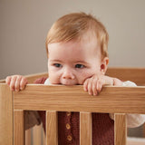 Baby in FLEXA OAK cot bed, safely biting top rail; child-safe design, oak finish, nursery setting visible.