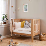 FLEXA BABY oak cot bed in a neutral nursery, soft bedding and child-safe rounded edges visible.